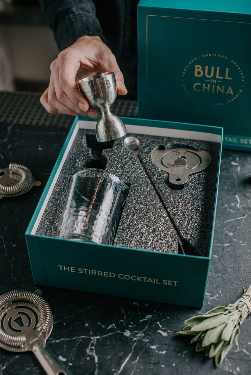 A hand holds a silver jigger above an open teal gift box containing a clear mixing glass, bar spoon, strainer, and bar tool set, with a branded Bull in China box behind it on a dark marble surface.